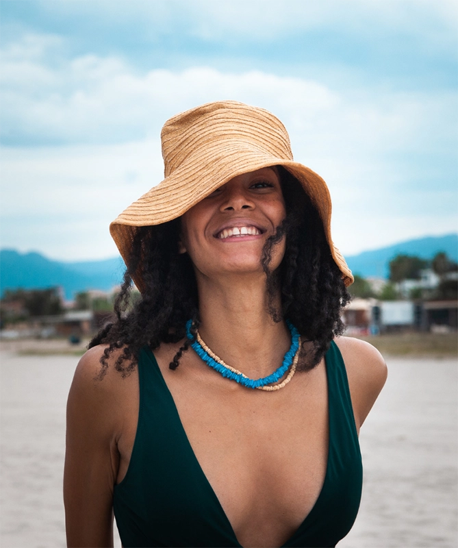A woman wearing a beige hat smiling as she stands on the beach