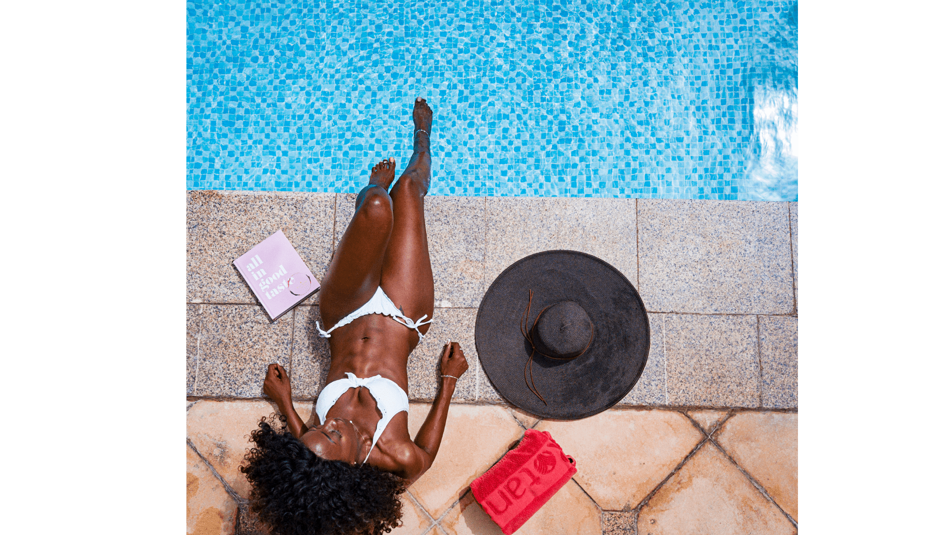 A woman sitting poolside with her feet dipped in the water with a book, hat and towel next to her
