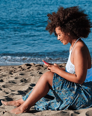 A woman sitting on the sand at the beach by the water while looking down and using her smartphone