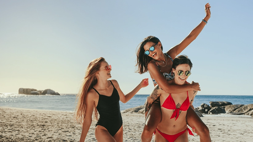 3 women friends walking down the beach in their swim gear with one woman carrying another on her back